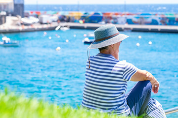 Fototapeta premium Rear view of senior caucasian woman sitting at the harbour admiring the sea. Mature lady with blue cap enjoying vacation looking the horizon over water