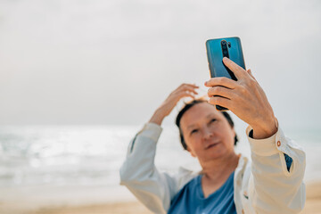 an adult woman on the beach near the ocean in the spring at a picnic takes a selfie, talks a video call, photographs the surrounding nature. Older generation and modern technologies