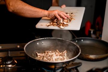 male chef pours chopped champignons from cutting boards into a frying pan