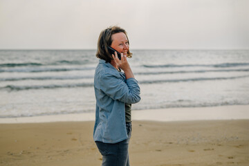 a young woman on the beach near the ocean in the spring at sunset talking on the phone with a smile on her face to her loved ones, friends to share the joy of vacation. modern technologies