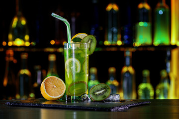Cocktail with kiwi, lemon, ice on bar counter in a restaurant, pub. Fresh prepared alcoholic cooler beverage at nightclub. Showcases with bottles on dark background