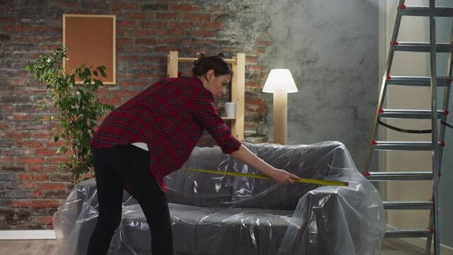 Woman measures sofa length covered with foil using tape in living room backside view. Lady works in room prepared for renovation. Home reconstruction