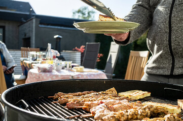 Woman with a plate and tongs for hot food stands near the barbecue with grill meat and cheese, in the courtyard of the house.