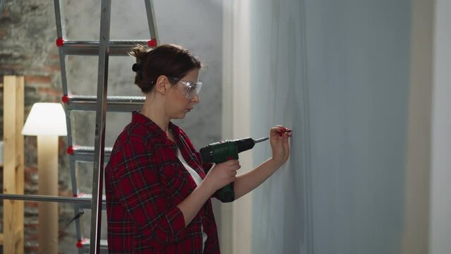 Woman with plastic glasses chooses place to screw fastening in wall making repair at home side view. Electric wireless screwdriver for easy renovation works