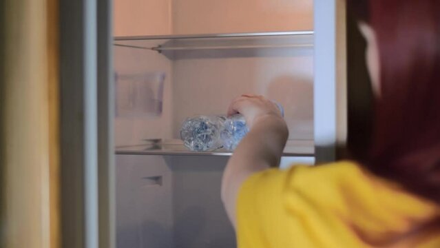 Mature adult woman with red hair is taking a bottle of cold water from the fridge and closing the fridge in the kitchen