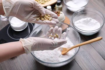 Fototapeta na wymiar Woman in gloves filling bath bomb mold with dried flower buds at wooden table, closeup