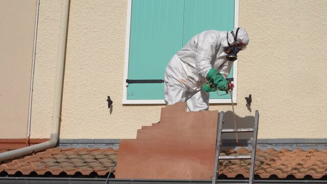 Man In PPE Suit Spray Painting The Tile Roof Of House. - wide