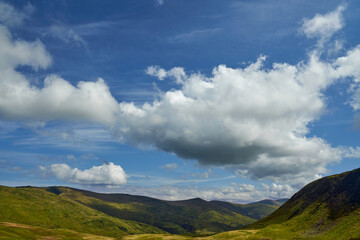 Fototapeta premium Huge cumulus clouds floating in blue sky, leaving shadows on grassy meadows outdoors. Panoramic view of sunlit valley between mountain hills in sunny day, with copy space. Concept of landscape. 