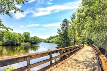Beneath the blue sky and white clouds of a beautiful summer day in Wisconsin, the Red Cedar State Bike Trail crosses a  wooden bridge and passes along the Red Cedar River through a lush green forest.