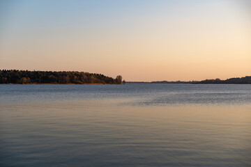 Lake landscape during sunset. Beautiful nature and calm water of the Lake Müritz. Reflections of the sky in the water. Trees and buildings are far away on the horizon.