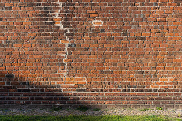 Old brick wall of a traditional building. A big crack in the house was repaired. Red bricks background texture. Abstract backdrop of an old facade with shadows and grass on the ground.