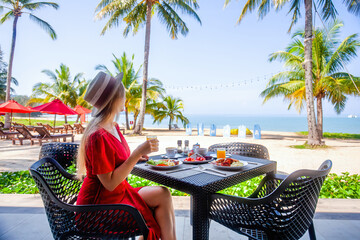 Woman on tropical healthy breakfast in hotel with sandy sea beach, palm trees and swing on background. Salad, fruits and fresh orange juice on morning food. Luxury resort, holidays in Thailand