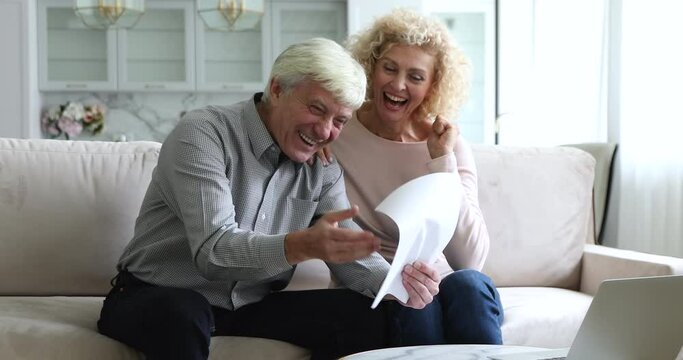 Happy mature spouses sit on couch reviewing received notification from bank about debt repayment, pension growth, read agreement with profitable terms smiling feel overjoyed by great news in document