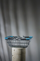 Mimi shopping cart or metallic basket with blue handles on concrete stand. Grey background with harsh shadows. Consumerism, market