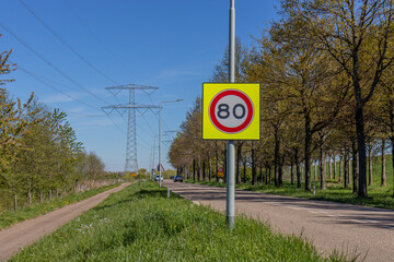 Traffic sign: maximum speed 80 between a rural road and a bicycle lane, trees, huge high voltage towers and circulating cars in the blurred background, sunny day in Netherlands