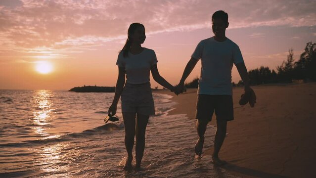 Happy Asian couple hands holding and walking together on the beach while beauty in nature of sunset time evening of vacation summer. Young in love romantic and relaxing at seaside. Honeymoon concept. 
