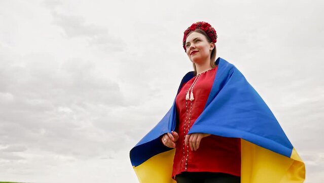 Smiling ukrainian woman with national flag on sky background. Portrait iof young lady in red embroidery Vyshyvanka and flowers wreath. Ukraine freedom, patriot symbol, victory in war.