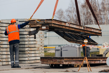 Slinger in helmet and vest controls unloading of metal structures on construction site. White handyman unloads load.