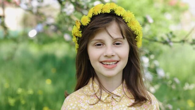 Little Girl with a wreath on her head Looking at Camera. Face Funny Contemplative Kid Child. Closeup smiling face with fresh spring flower in sunlight. Face of happy girl enjoying flower buds