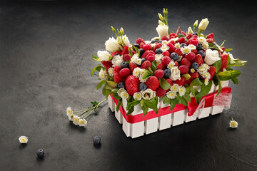 White wooden box filled with flowers, strawberries and raspberries, decorated with mint leaves on a black background