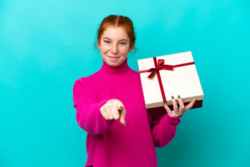 Young caucasian reddish woman holding a gift isolated on blue background points finger at you with a confident expression