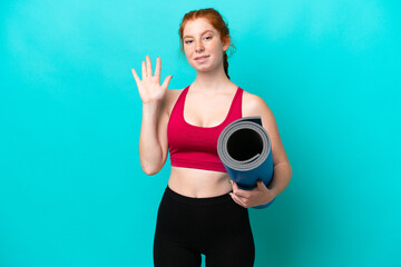 Young sport reddish woman going to yoga classes while holding a mat isolated on blue background saluting with hand with happy expression