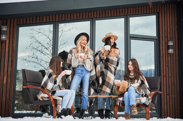 Young women enjoying winter weekends on terrace of contemporary barnhouse in the Scandinavian style. Four girls having fun, sitting on chairs and drinking hot tea.