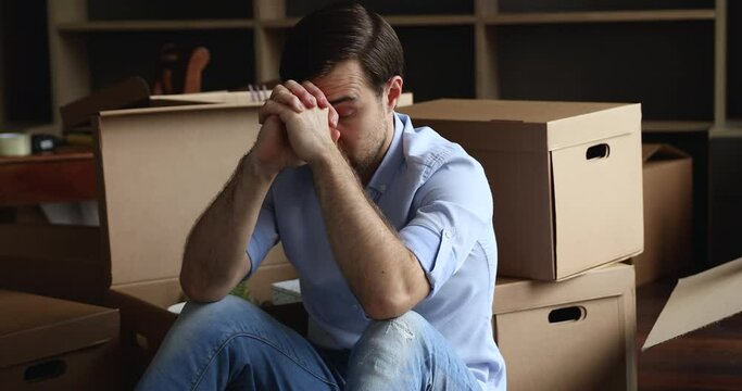 Exhausted man sit on floor near heap of boxes with stuff looks depressed relocate to new flat due to divorce or break up, feels unmotivated to unpack belongings. Financial problems, eviction concept