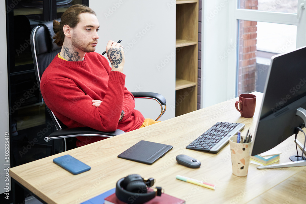 Poster Young graphic designer sitting at desk and looking at computer monitor with pensive expression during his work at office