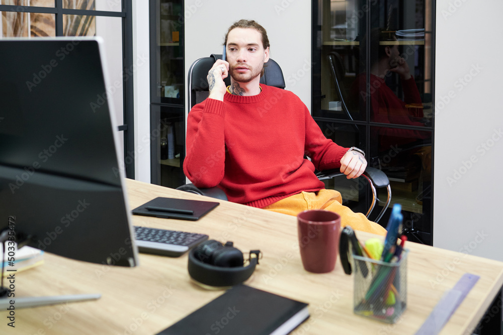 Poster young businessman sitting on chair in front of his workplace with computer and talking on mobile pho