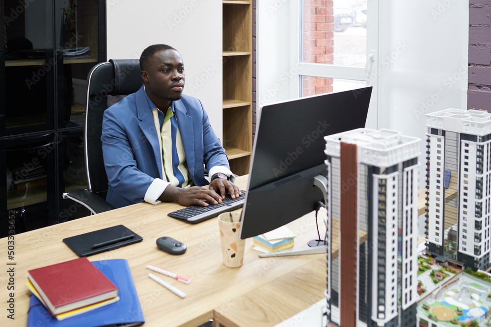 Poster African serious architect looking at monitor while typing on computer at his workplace at office with models of buildings on table