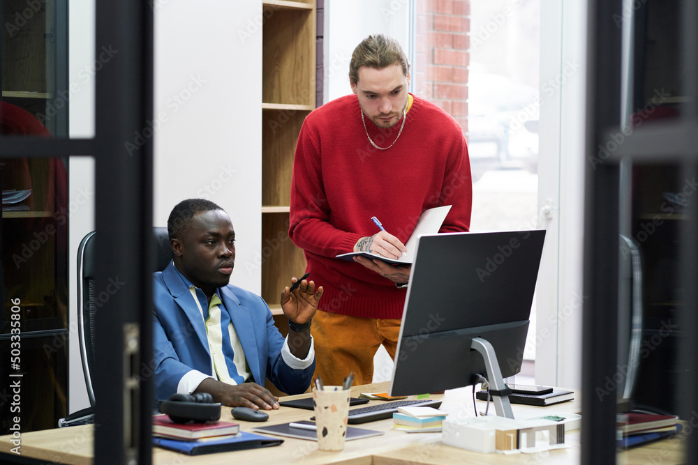 Poster African manager sitting at his workplace with computer and giving work to businessman while he making notes in his notebook