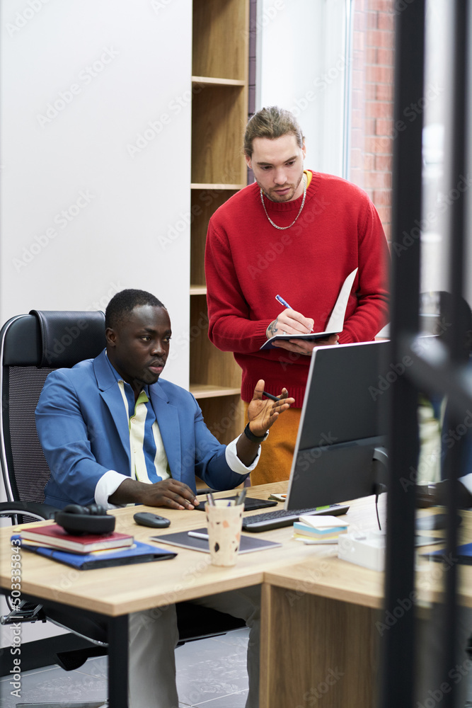 Poster Young businessman making notes in notebook while director sitting at table and pointing at monitor explaining him the work