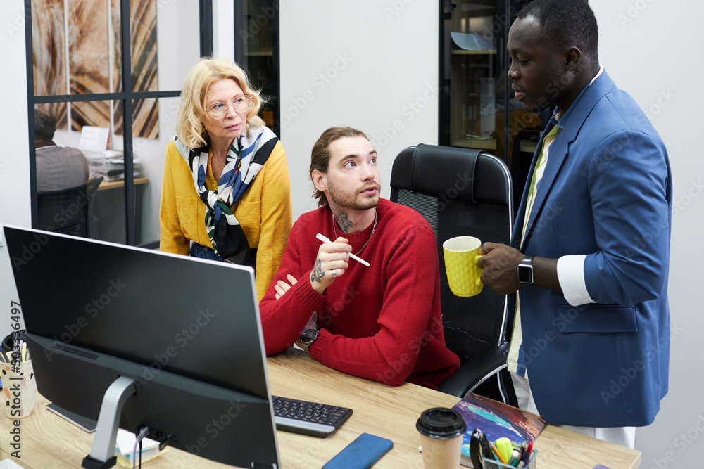 Poster graphic designer sitting at his workplace with computer and talking to his colleagues, they working 