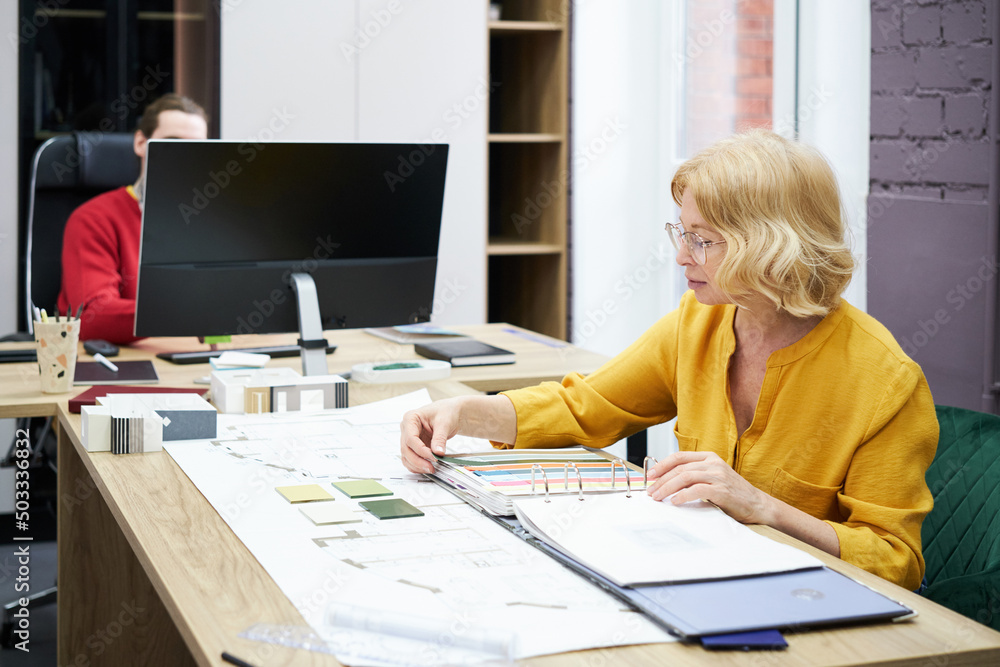 Poster Designer deciding colour samples at table with her colleague working on computer in background