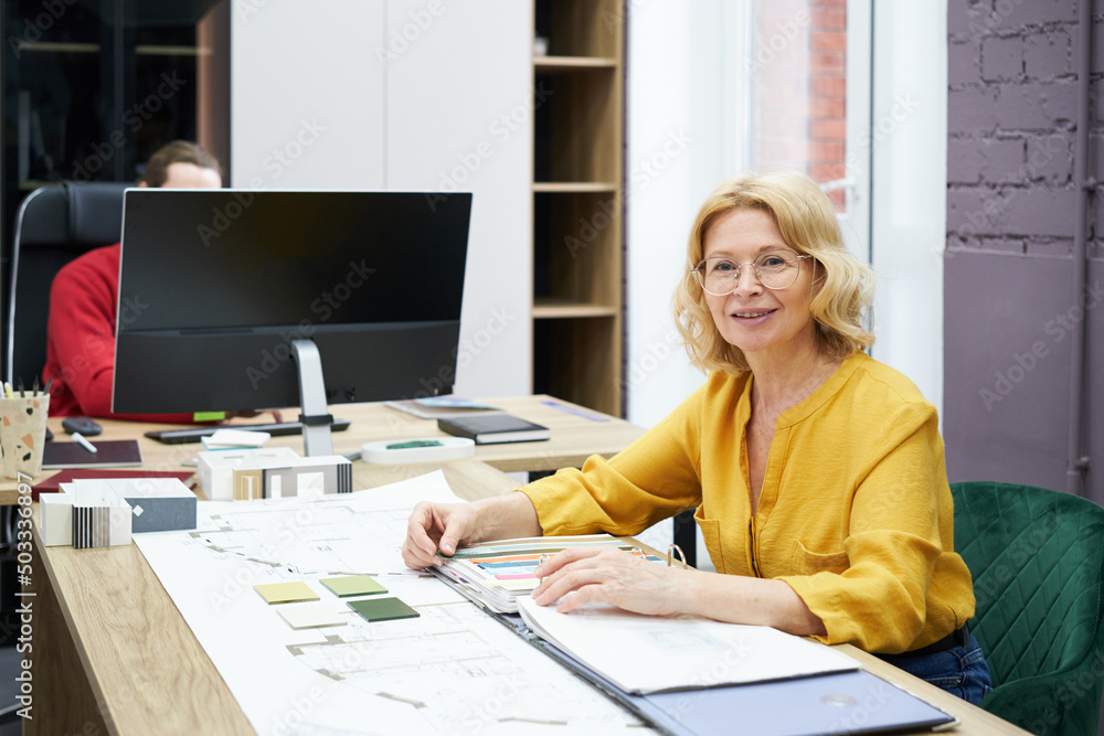 Poster Portrait of mature designer smiling at camera while working at table with colour samples