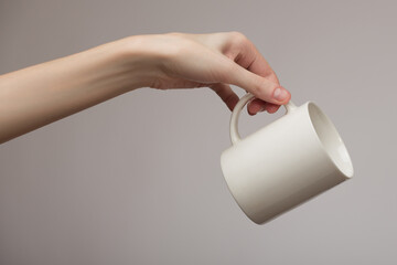 A woman's hand gracefully holds an empty white mug. Gray background.