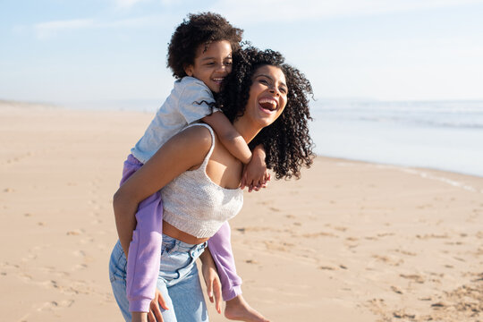 Contented mother and daughter spending time on beach. African American family walking, laughing, playing, riding on back. Leisure, family time, parenthood concept