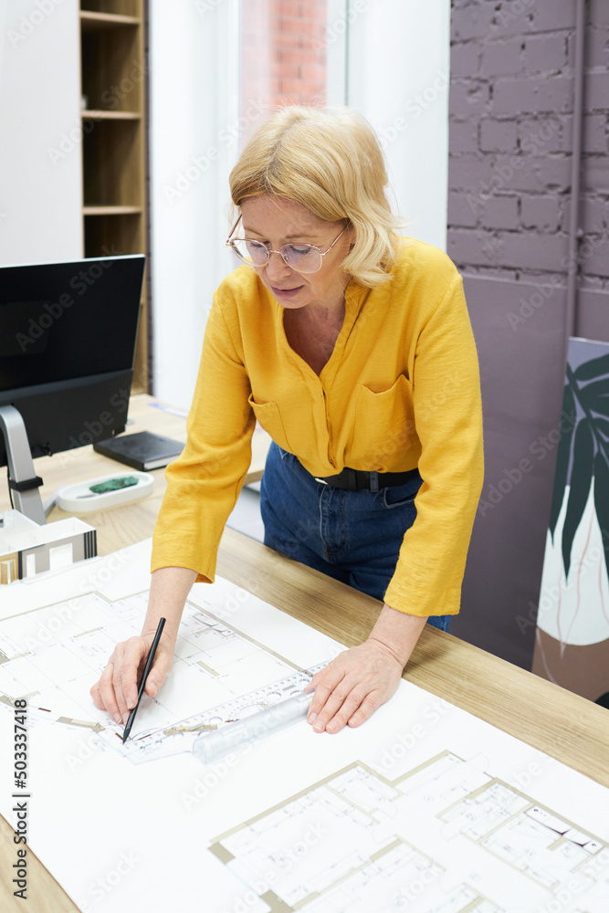 Poster Serious female engineer in eyeglasses working with blueprint on table using pencil and ruler