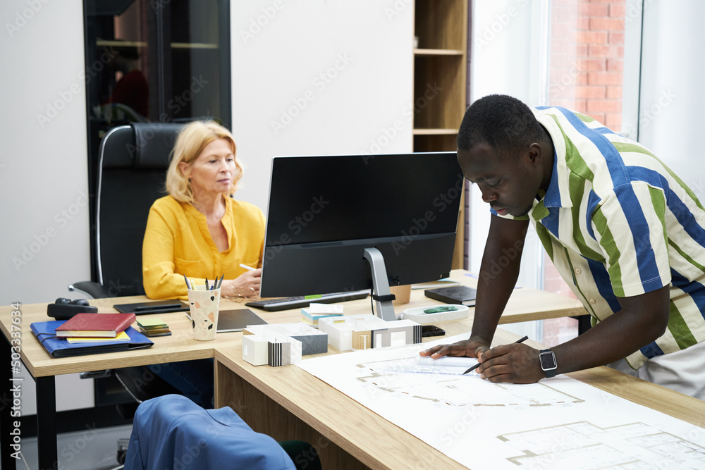 Poster African male engineer drawing blueprint at table with his colleague working on computer in background