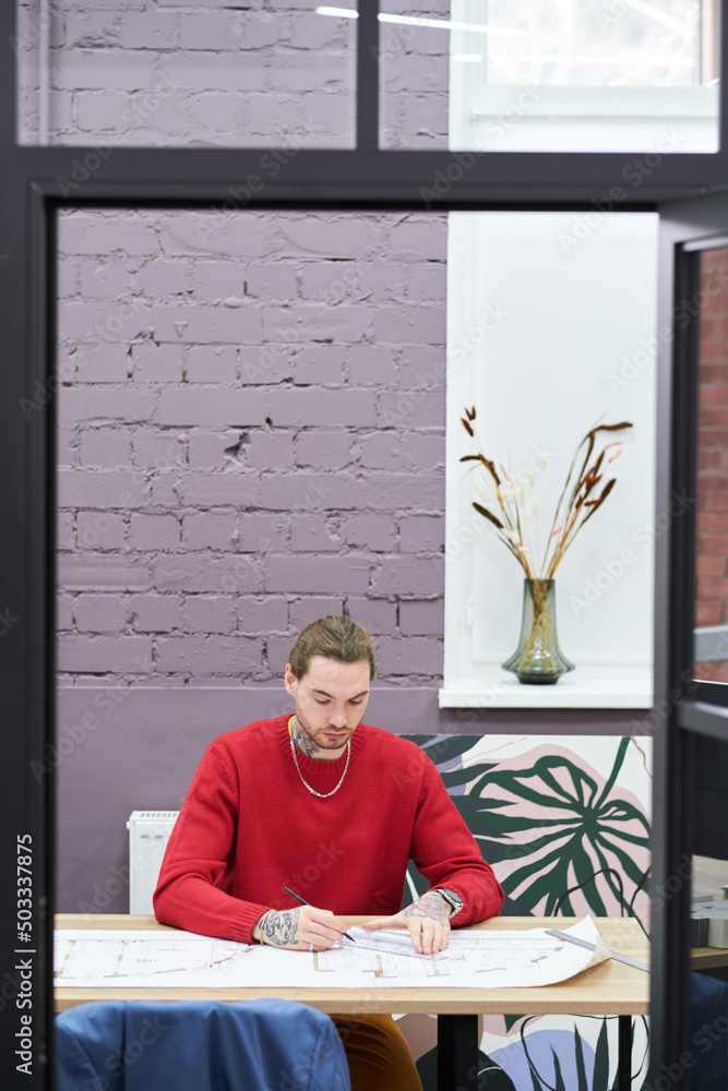 Poster Young architect using ruler to draw blueprint of new construction project while sitting at workplace at office