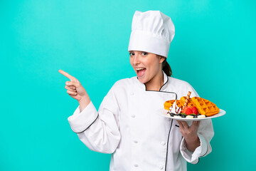 Young caucasian chef woman holding waffles isolated on blue background surprised and pointing finger to the side