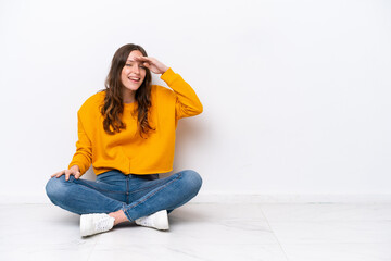 Young caucasian woman sitting on the floor isolated on white wall looking far away with hand to look something