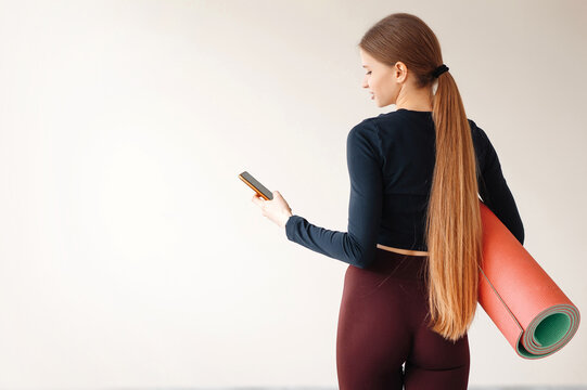 Woman with ponytail standing at work out or yoga class with rolled fitness mat and smartphone
