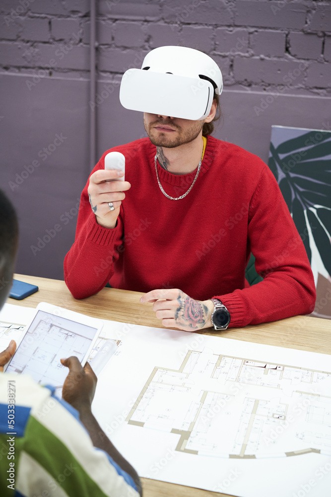 Poster Young designer sitting at table with blueprint and using virtual reality headset to look at new project