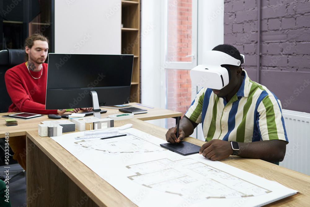Poster African designer in virtual reality goggles using graphic tablet at table with blueprint during work at office