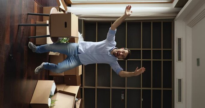 Young man dance to music enjoy relocation day, celebrate life changes in cozy unfurnished living room with heap of cardboard boxes full of personal stuff, vertical view. Move-in, bank mortgage concept