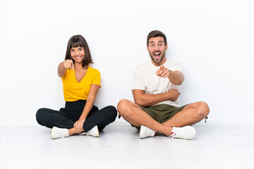 Fototapeta na wymiar Young couple sitting on the floor isolated on white background points finger at you with a confident expression