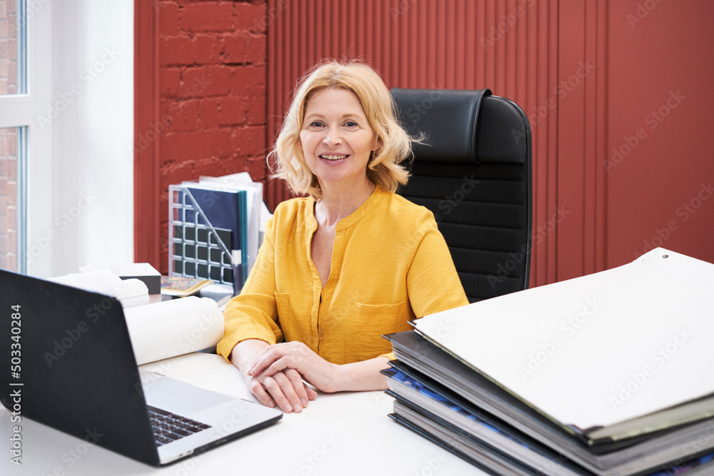Poster portrait of mature businesswoman smiling at camera while sitting at desk with laptop and stack of fo