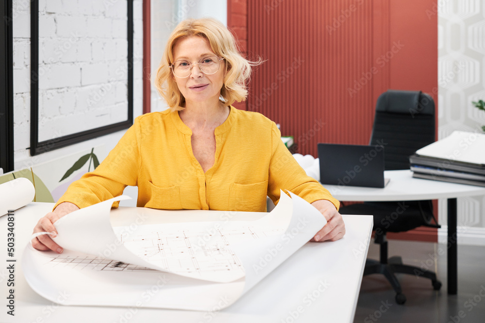 Aufkleber portrait of mature female architect in eyeglasses looking at camera while working with blueprints of